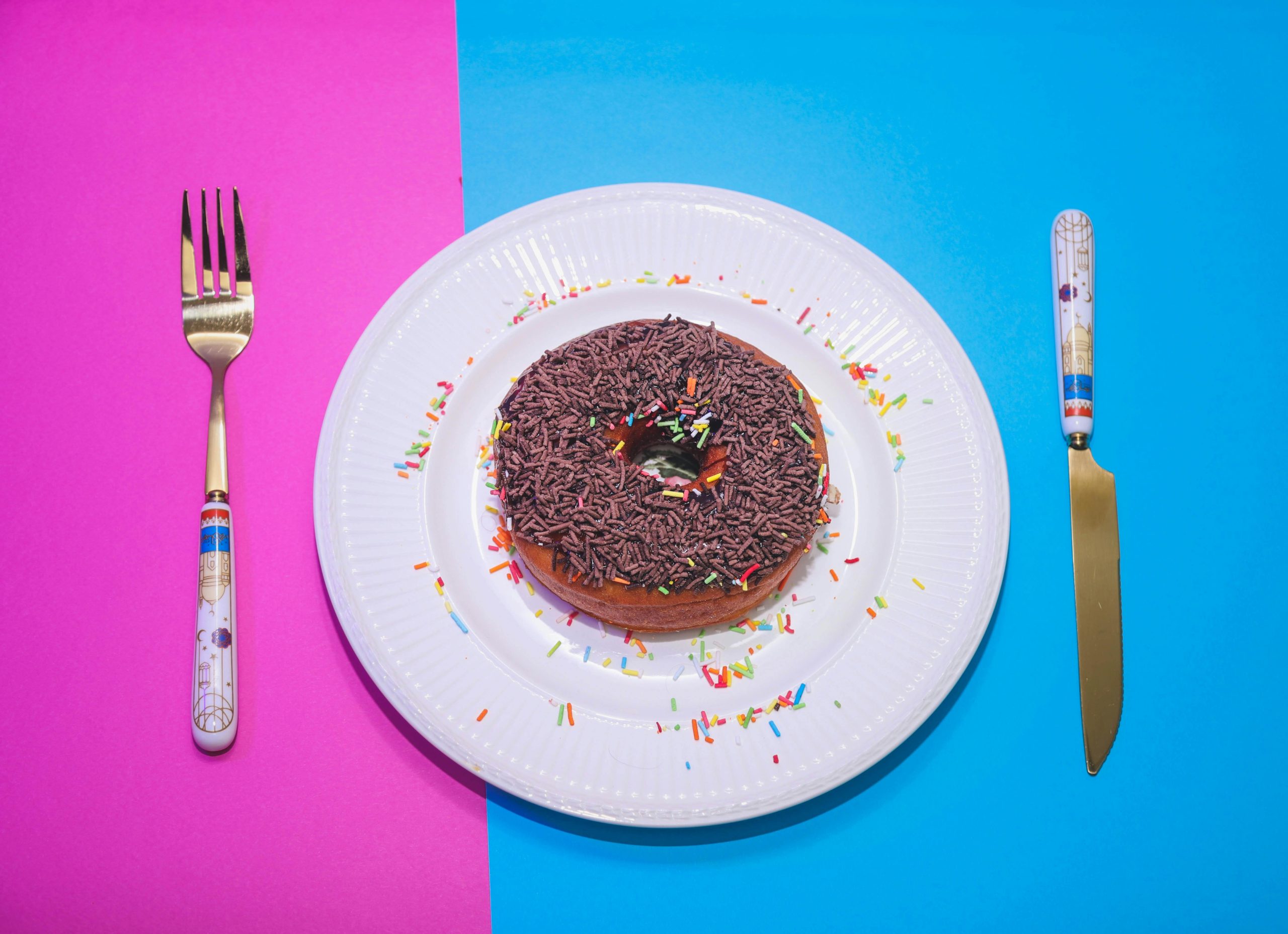 Colorful chocolate donut with sprinkles on a white plate, styled with cutlery.