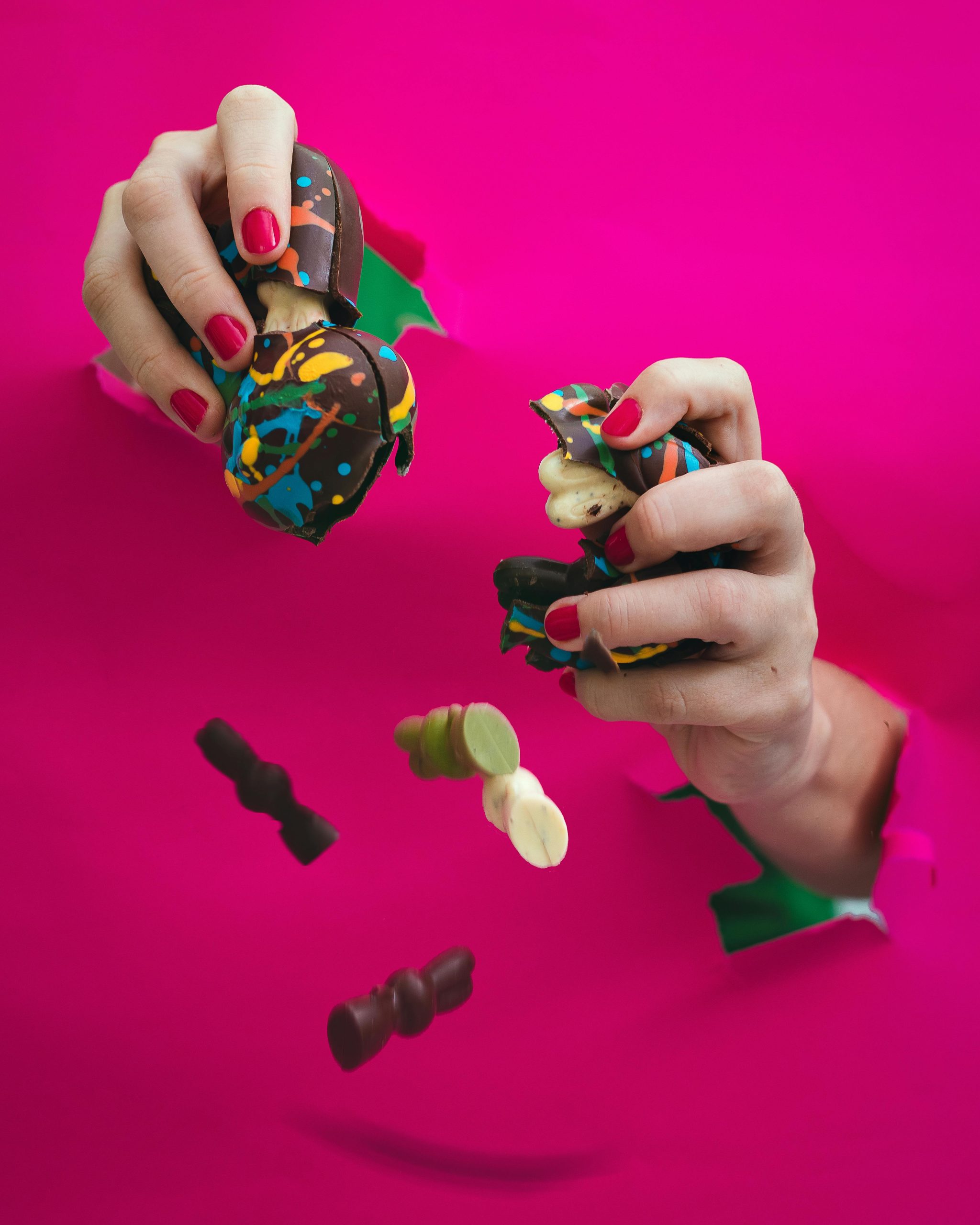 Hands breaking colorful chocolate candies against a vivid pink backdrop, showcasing creativity and sweetness.